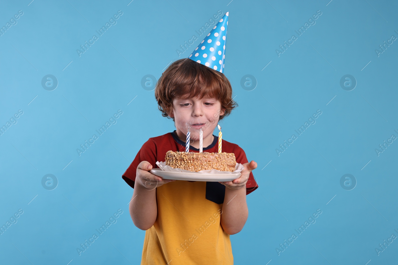 Photo of Birthday celebration. Cute little boy in party hat holding tasty cake with burning candles on light blue background