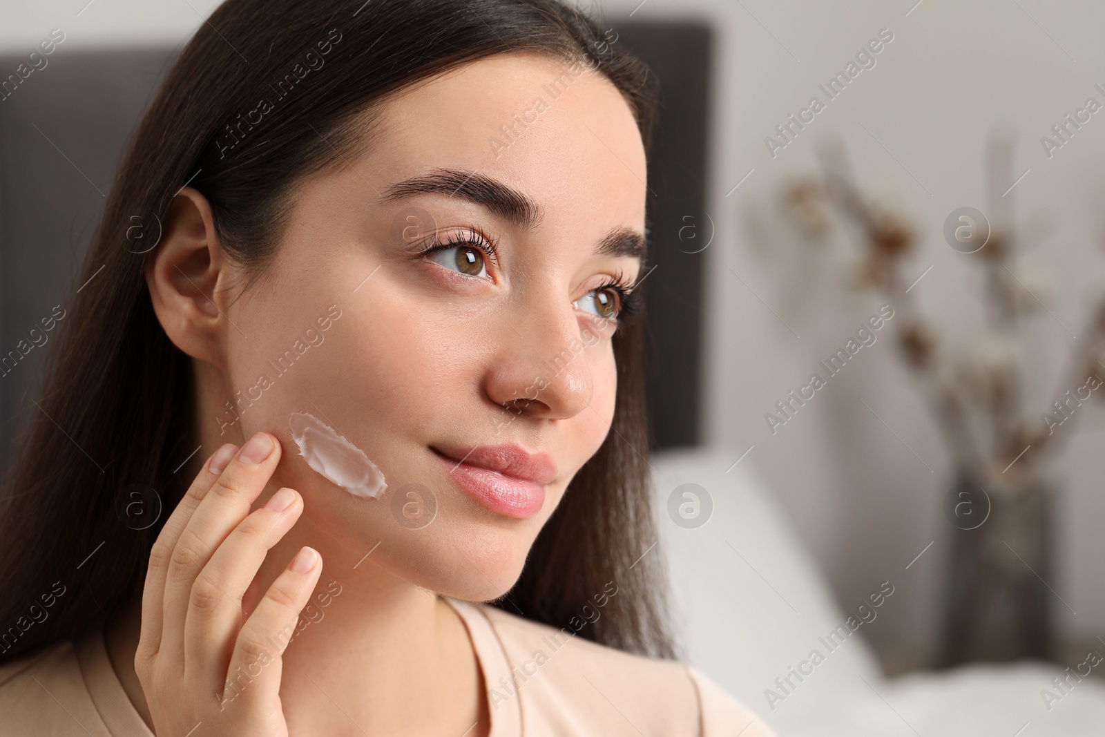 Photo of Young woman with dry skin applying cream onto her face indoors, closeup. Space for text