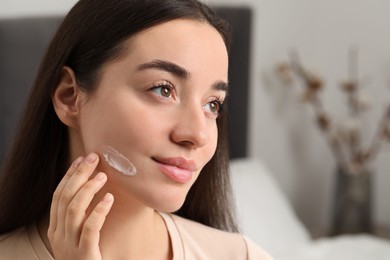 Photo of Young woman with dry skin applying cream onto her face indoors, closeup. Space for text