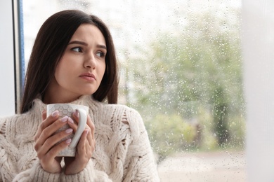 Photo of Sad beautiful woman with cup near window indoors on rainy day