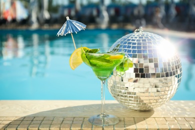 Photo of Shiny disco ball and refreshing cocktail on edge of swimming pool. Party items