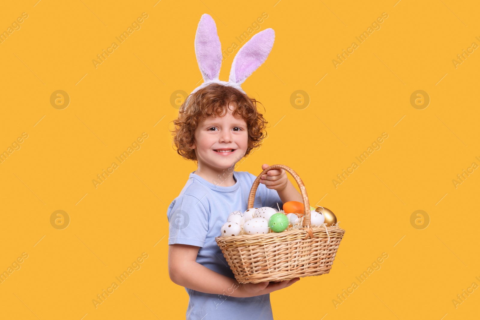 Photo of Portrait of happy boy in cute bunny ears headband holding wicker basket with Easter eggs on orange background