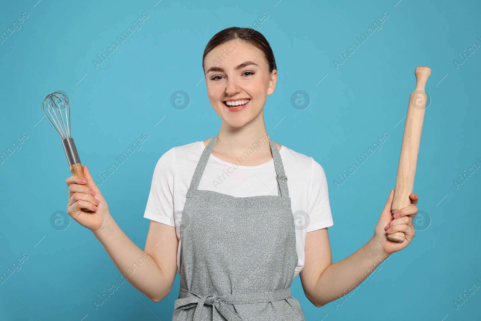 Photo of Beautiful young woman in clean apron with kitchen tools on light blue background