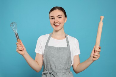 Beautiful young woman in clean apron with kitchen tools on light blue background