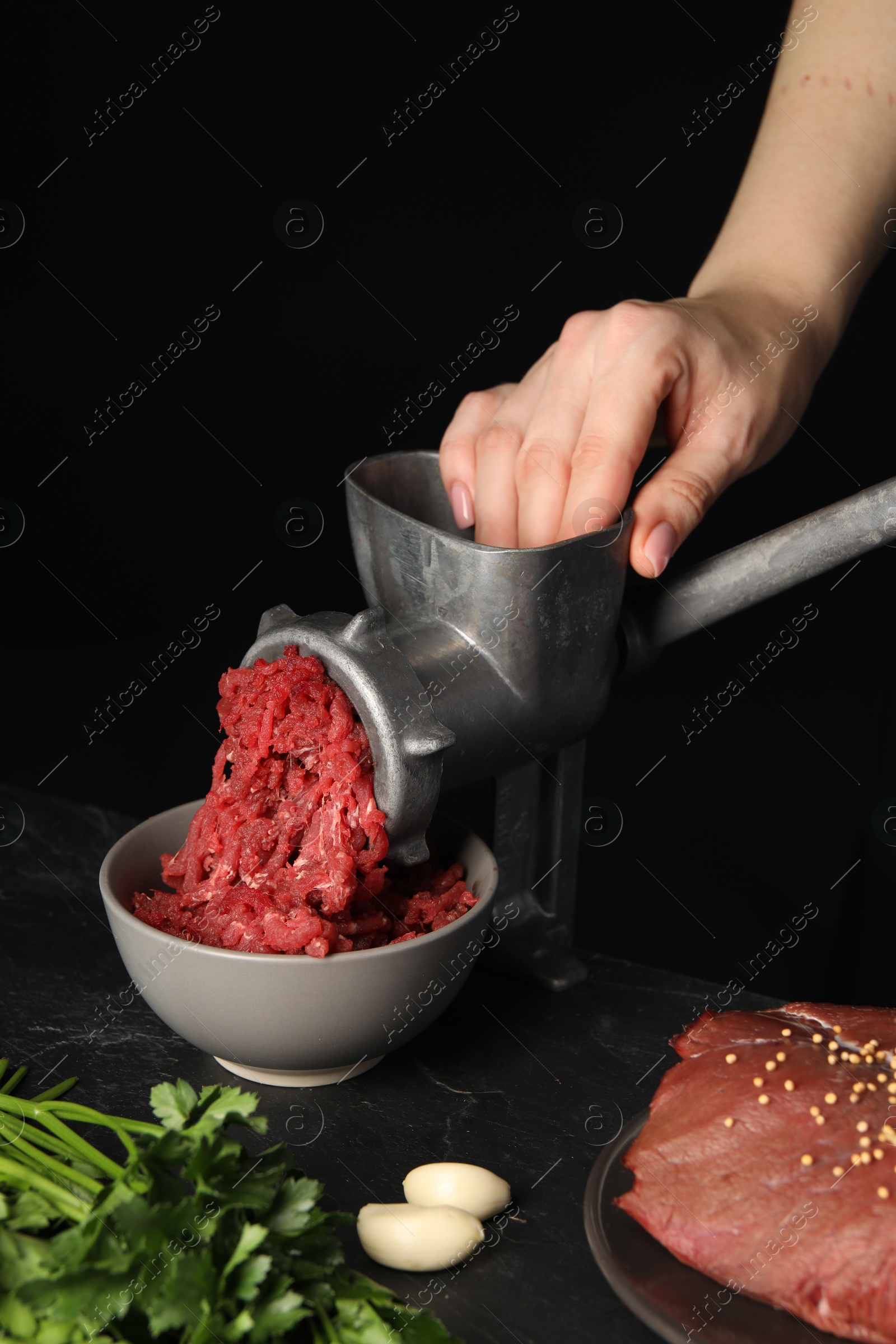 Photo of Woman making beef mince with manual meat grinder at table against black background, closeup