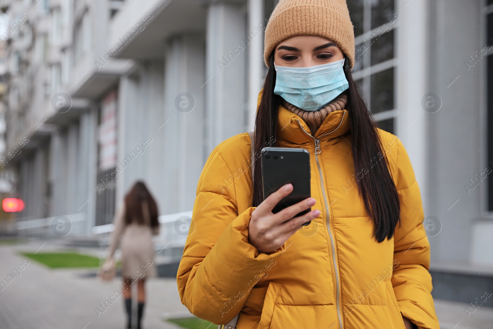 Photo of Young woman in medical face mask with smartphone walking outdoors. Personal protection during COVID-19 pandemic