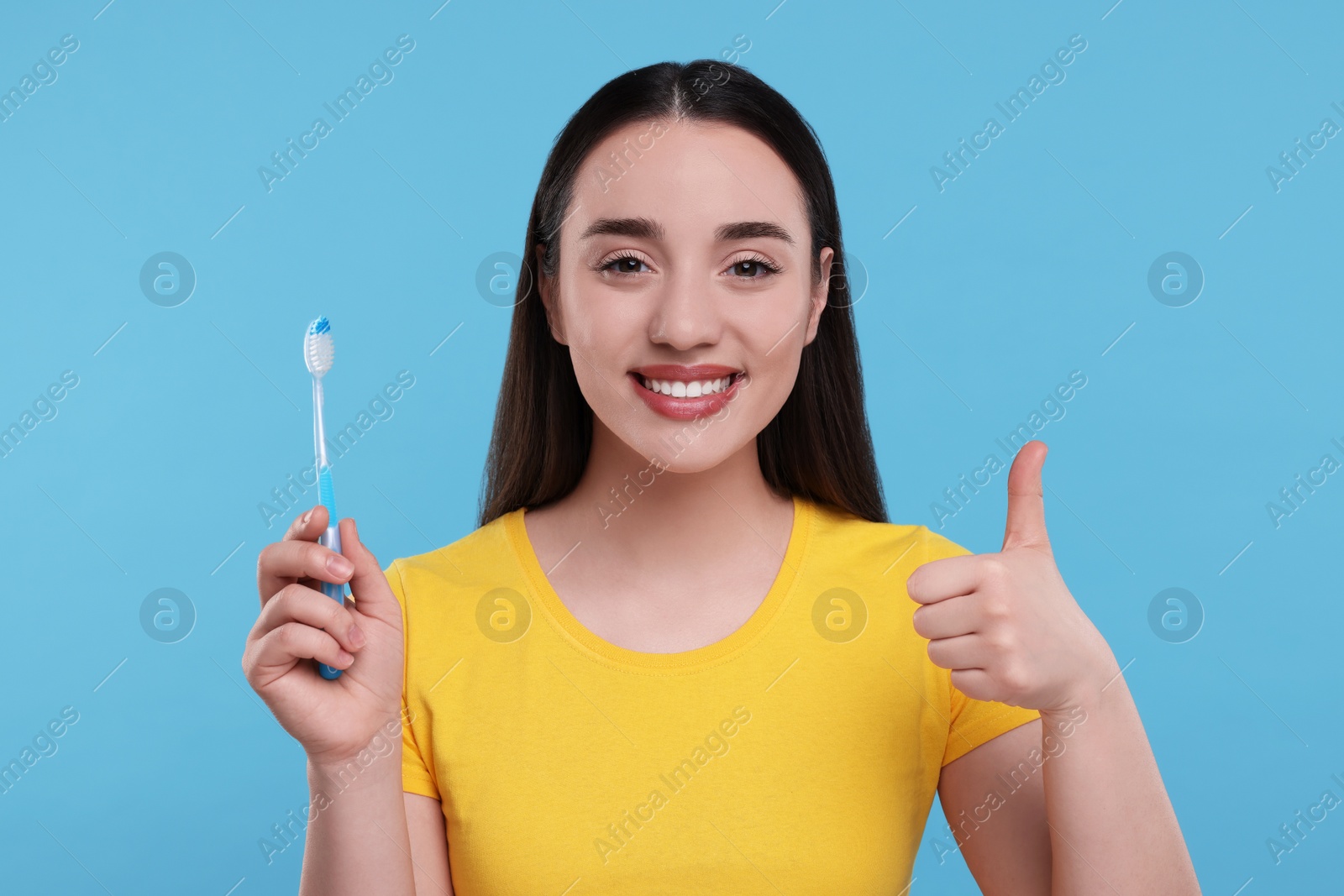 Photo of Happy young woman holding plastic toothbrush and showing thumb up on light blue background