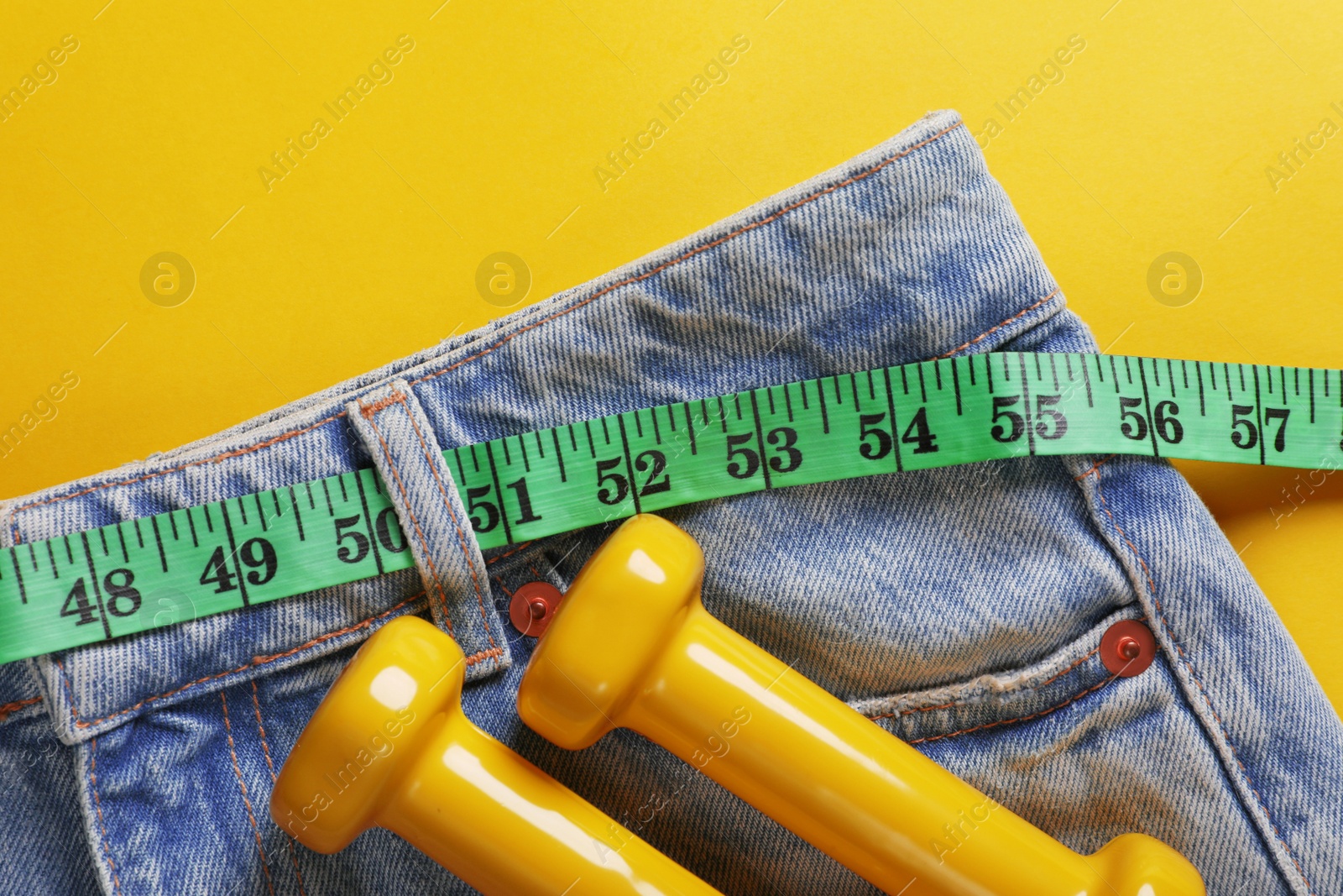 Photo of Jeans with measuring tape and dumbbells on yellow background, top view. Weight loss concept