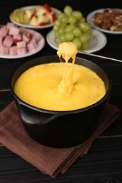 Dipping piece of ham into fondue pot with melted cheese on black wooden table, closeup