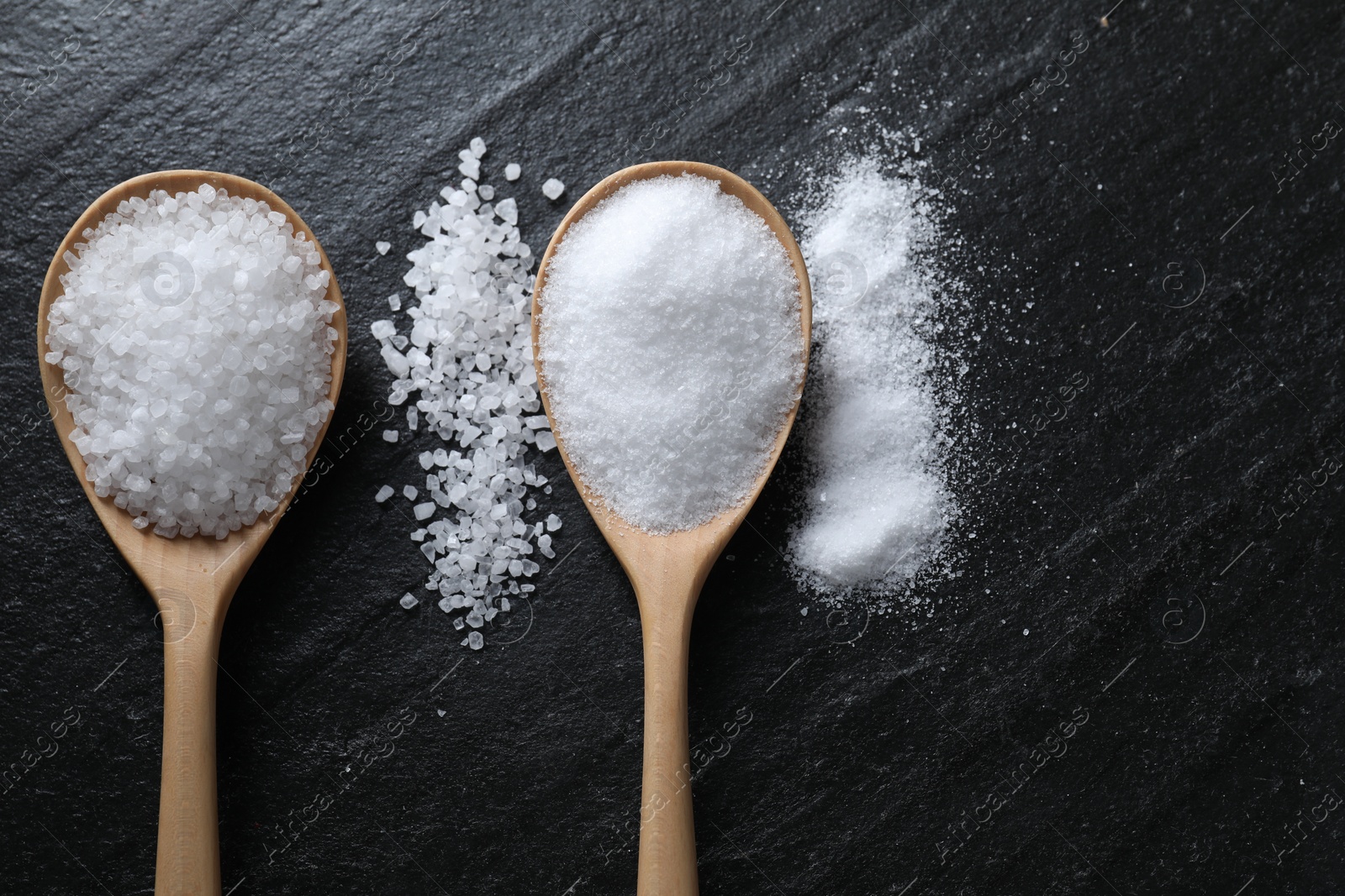 Photo of Organic salt in spoons on black table, flat lay. Space for text