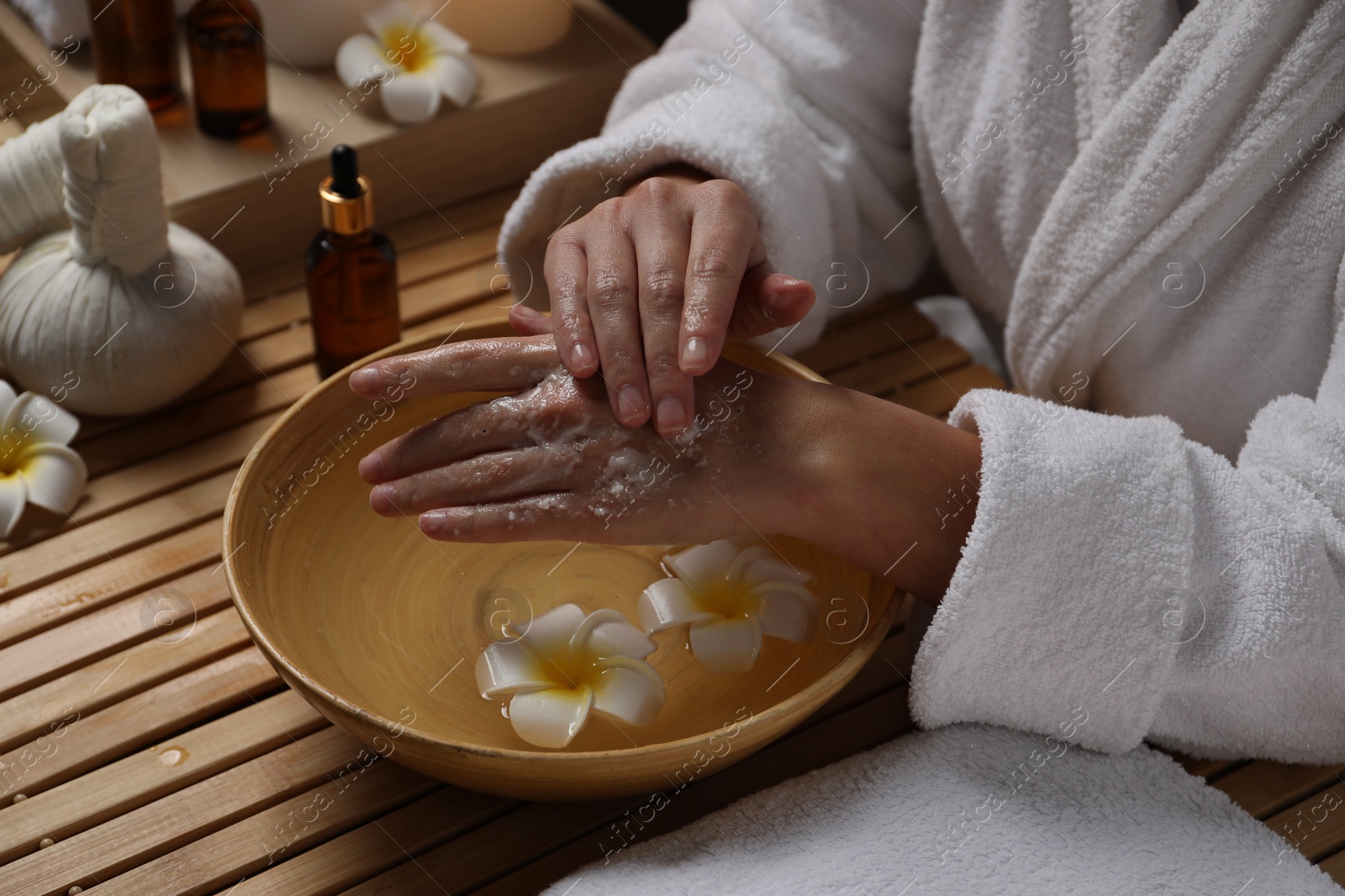 Photo of Woman applying scrub onto her hands in spa, closeup. Bowl of water and flowers on wooden table