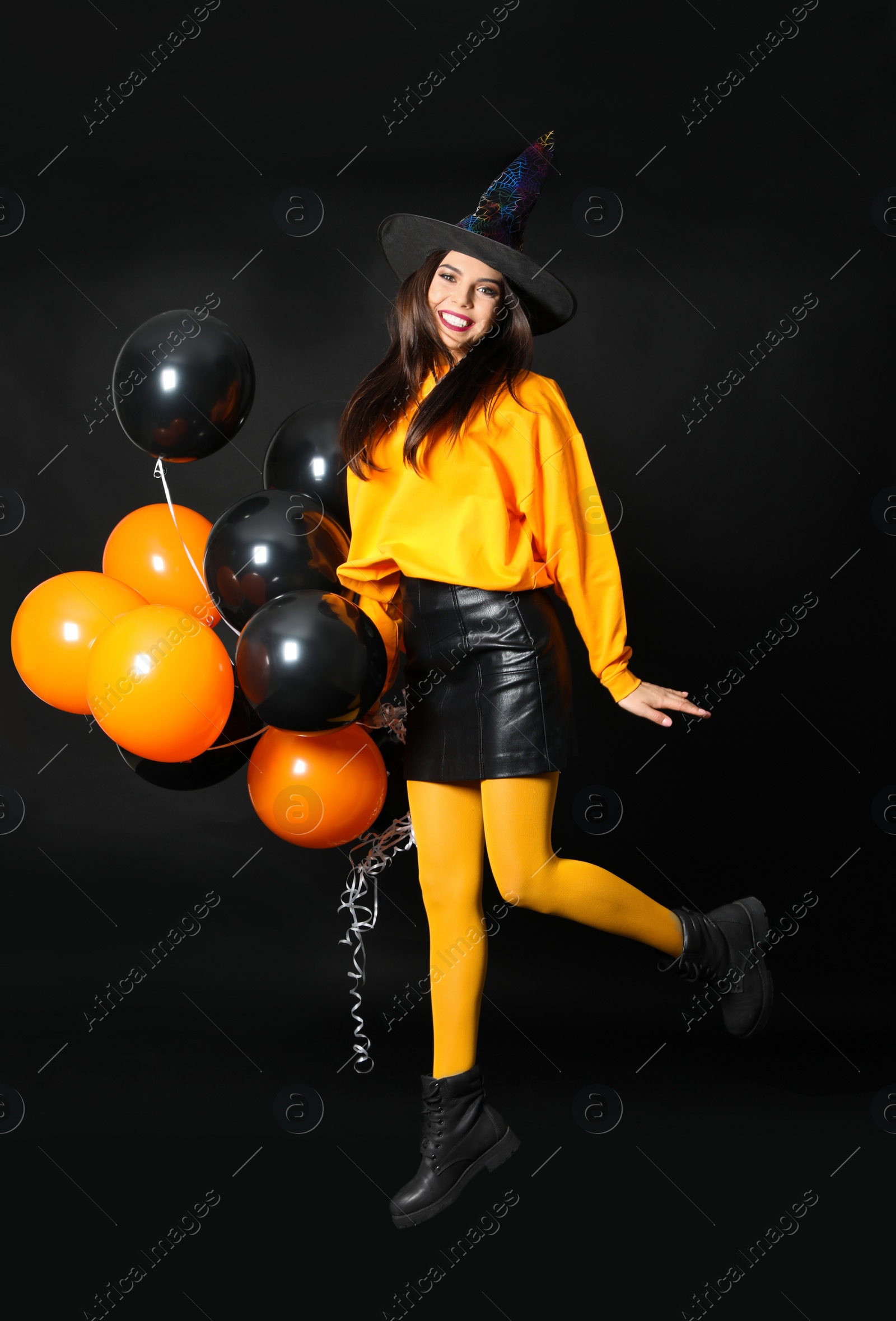 Photo of Beautiful woman wearing witch costume for Halloween party jumping with balloons on black background