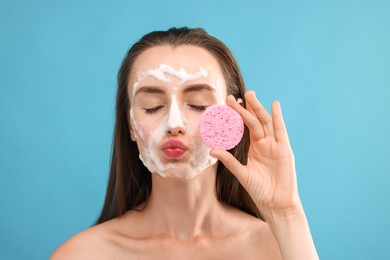 Happy young woman washing her face with sponge on light blue background