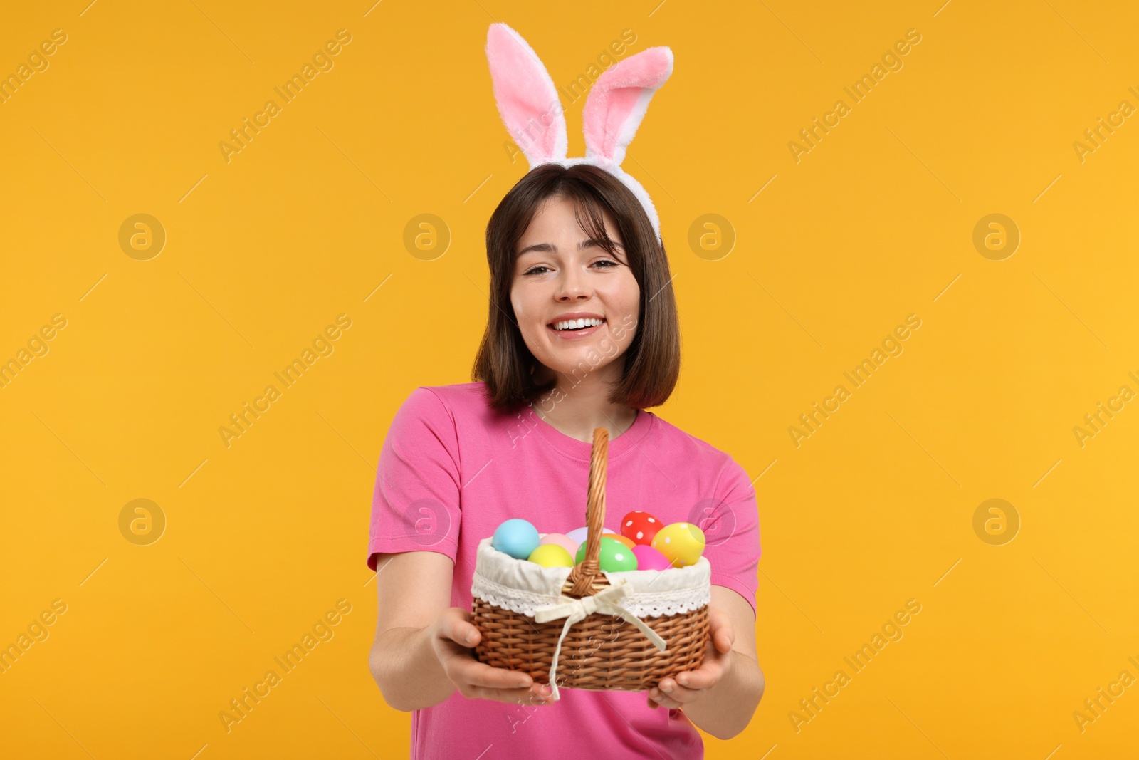 Photo of Easter celebration. Happy woman with bunny ears and wicker basket full of painted eggs on orange background