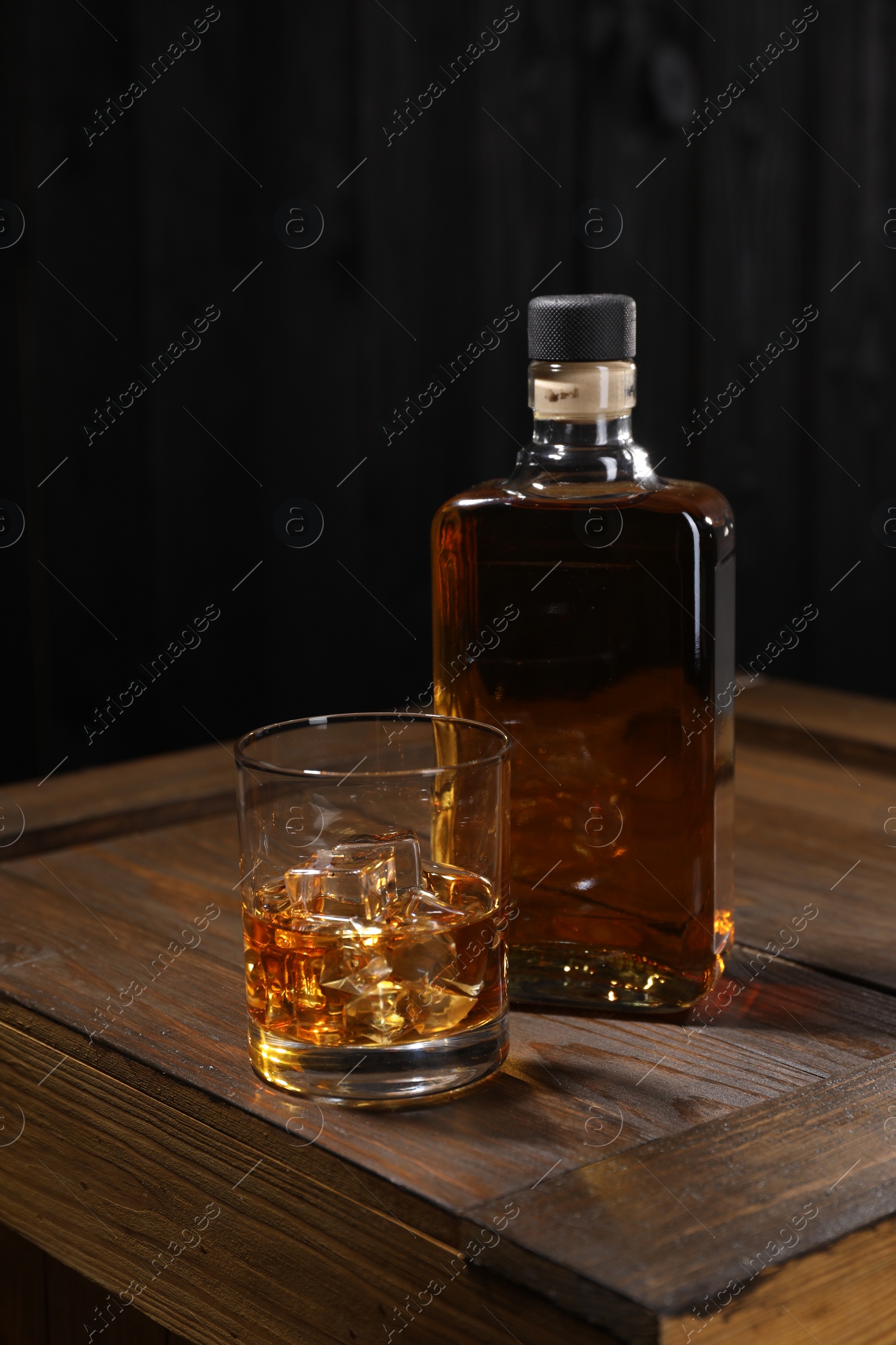 Photo of Whiskey with ice cubes in glass and bottle on wooden crate against black background