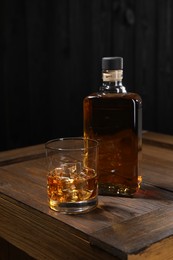 Whiskey with ice cubes in glass and bottle on wooden crate against black background