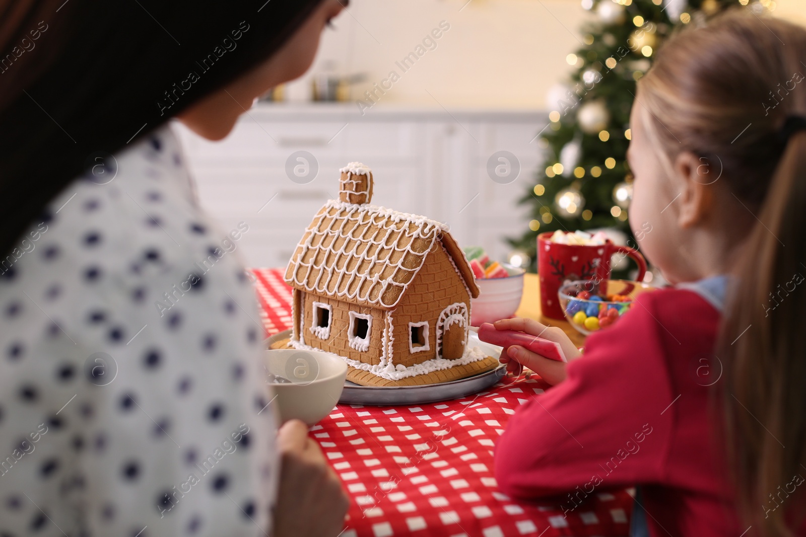 Photo of Mother and daughter decorating gingerbread house at table indoors