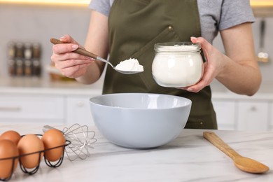 Housewife adding flour into bowl at white marble table in kitchen, closeup. Cooking process