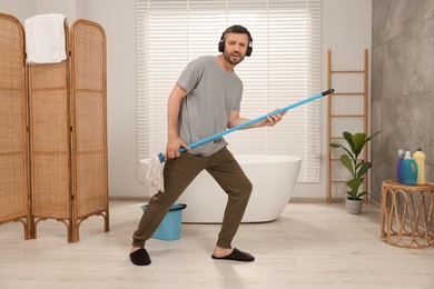 Photo of Enjoying cleaning. Happy man in headphones with mop listening to music while tidying up in bathroom