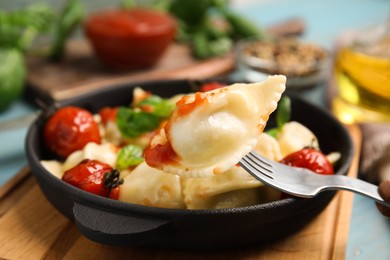 Fork with tasty ravioli and tomato sauce above pan on wooden board, closeup