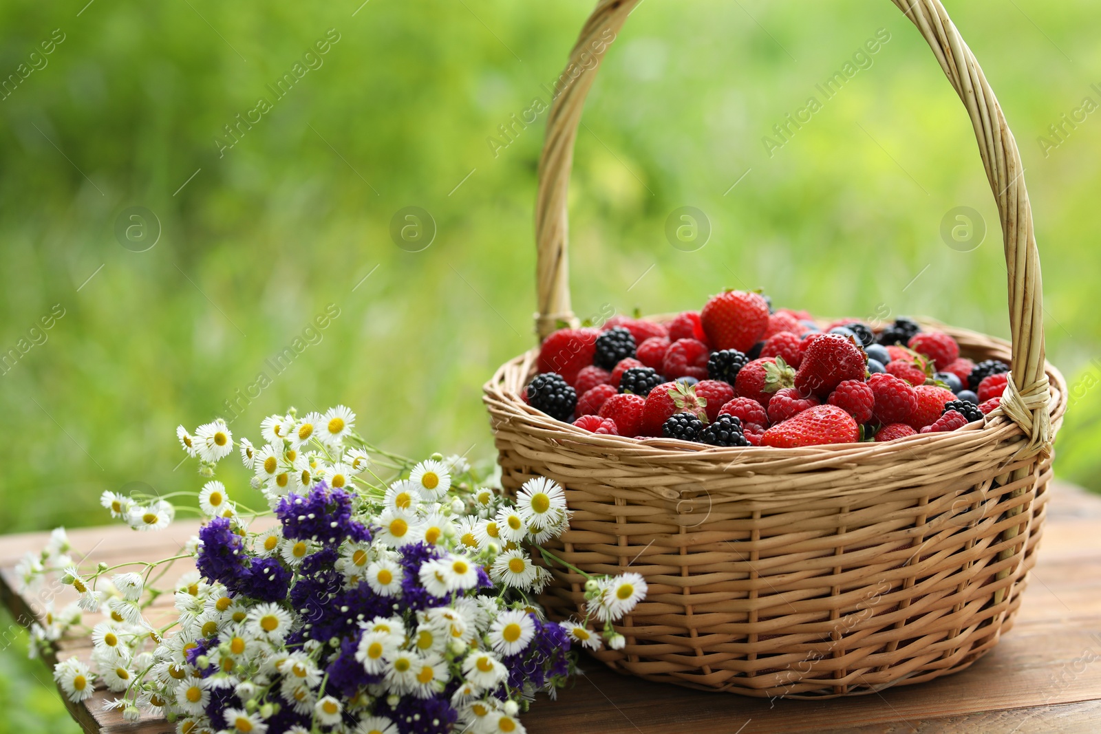 Photo of Wicker basket with different fresh ripe berries and beautiful flowers on wooden table outdoors