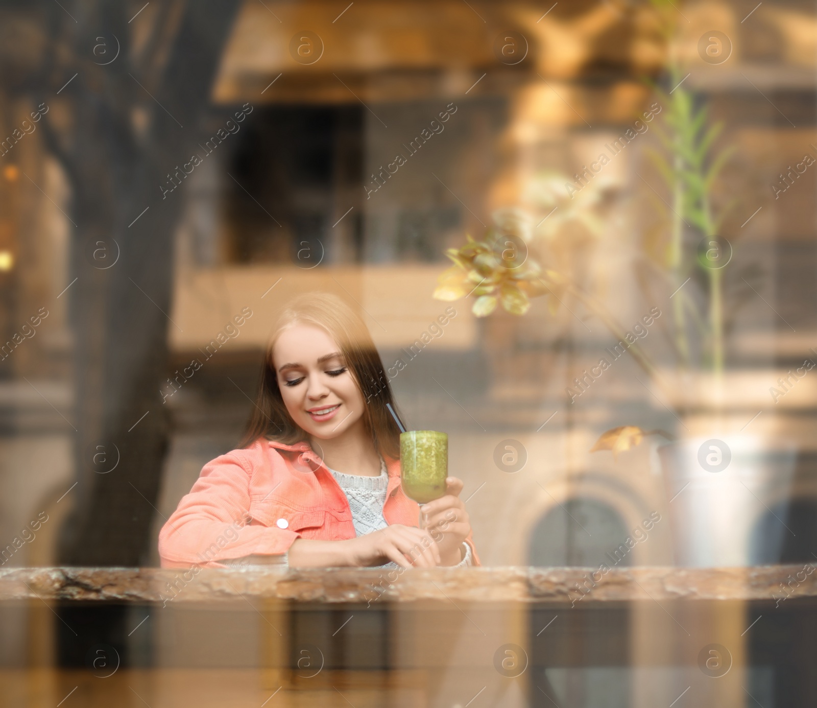 Photo of Pretty young woman with cocktail at table in cafe, view from outdoors through window