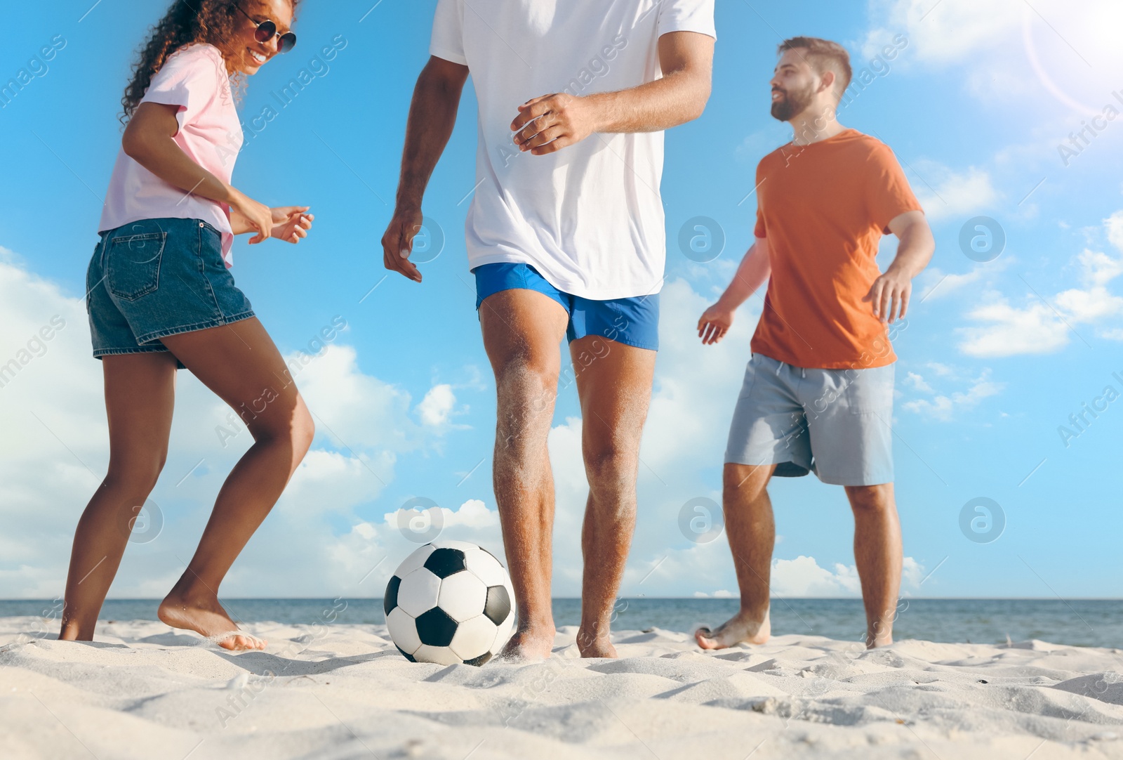 Image of Happy friends playing football on beach during sunny day, low angle view