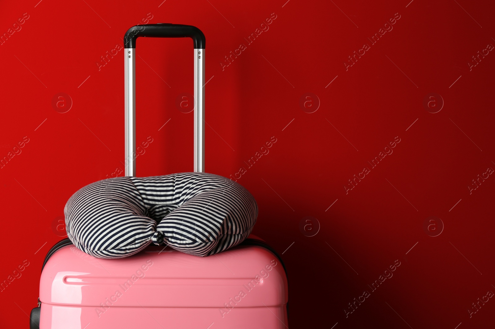 Photo of Pink suitcase with striped travel pillow on red background, space for text