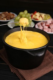 Dipping piece of ham into fondue pot with melted cheese on black wooden table, closeup