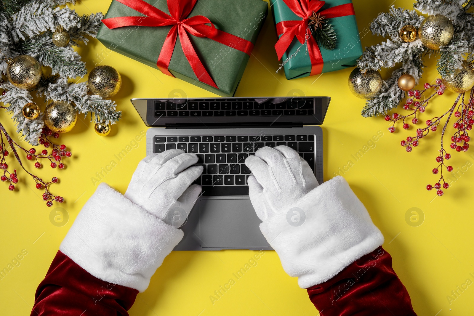 Photo of Santa Claus using laptop, closeup. Gift boxes and Christmas decor on yellow background, top view