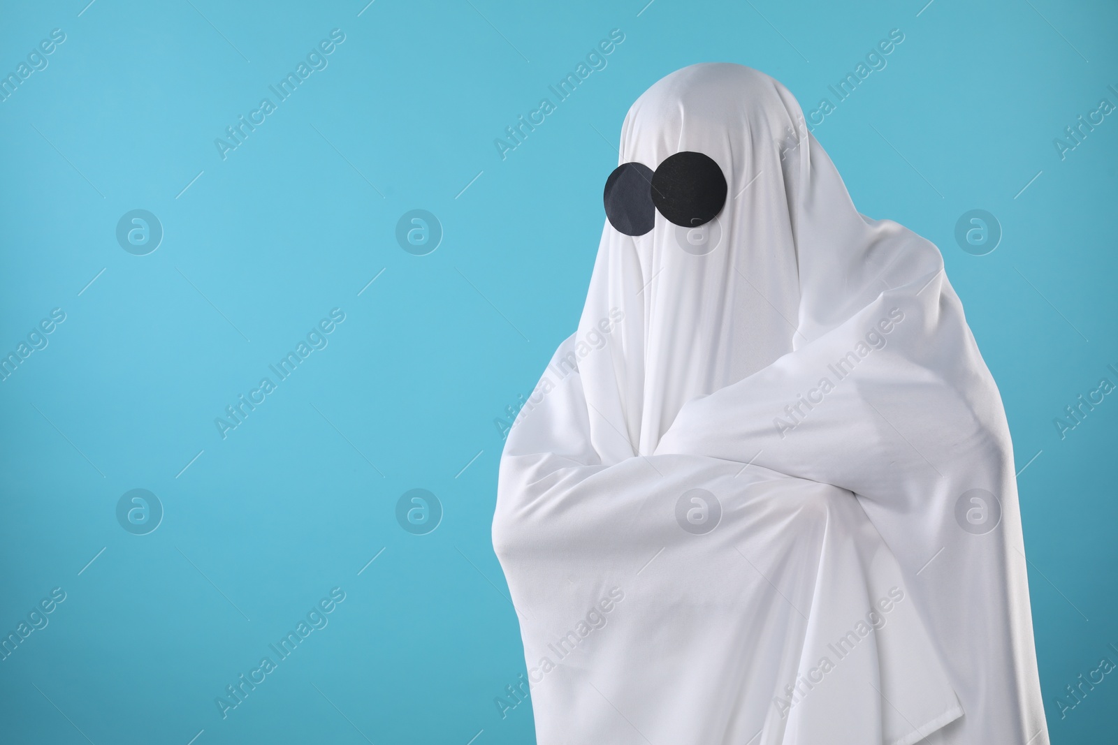 Photo of Creepy ghost. Person covered with white sheet on light blue background, space for text