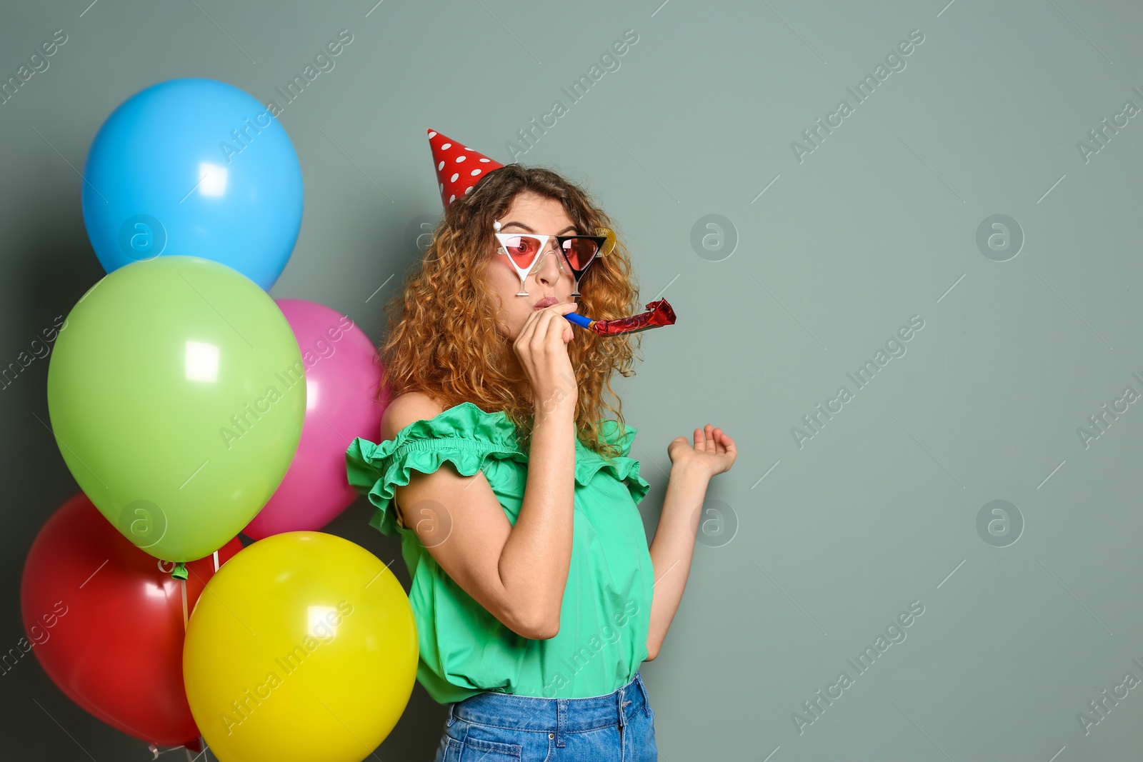 Photo of Young woman with party blower near bright balloons on color background. Birthday celebration