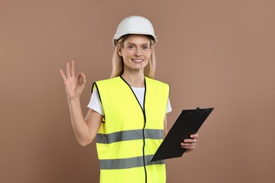 Photo of Engineer in hard hat holding clipboard and showing ok gesture on brown background