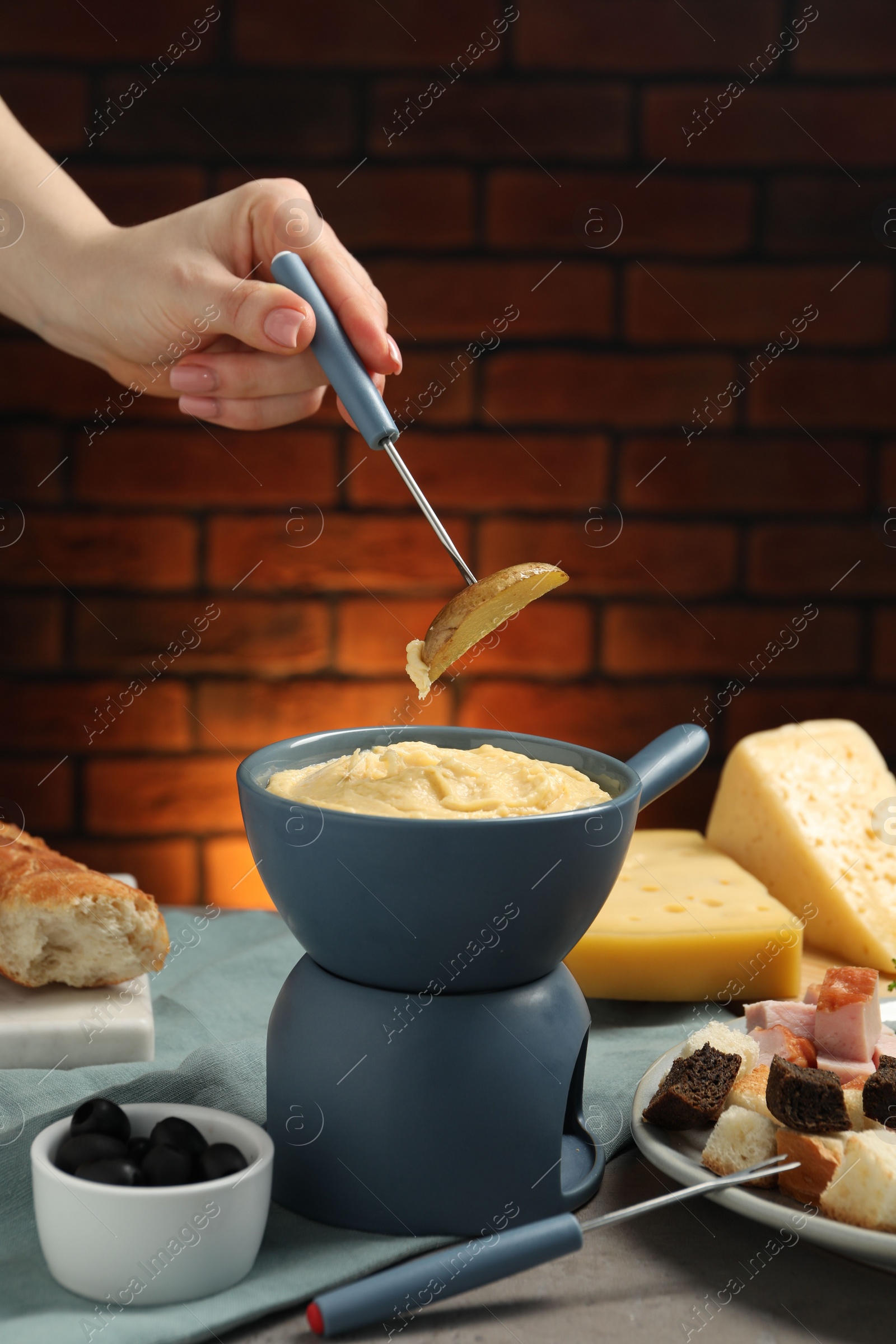 Photo of Woman dipping piece of potato into fondue pot with melted cheese at table with snacks, closeup