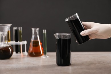 Woman pouring black crude oil into beaker at grey table against dark background, closeup
