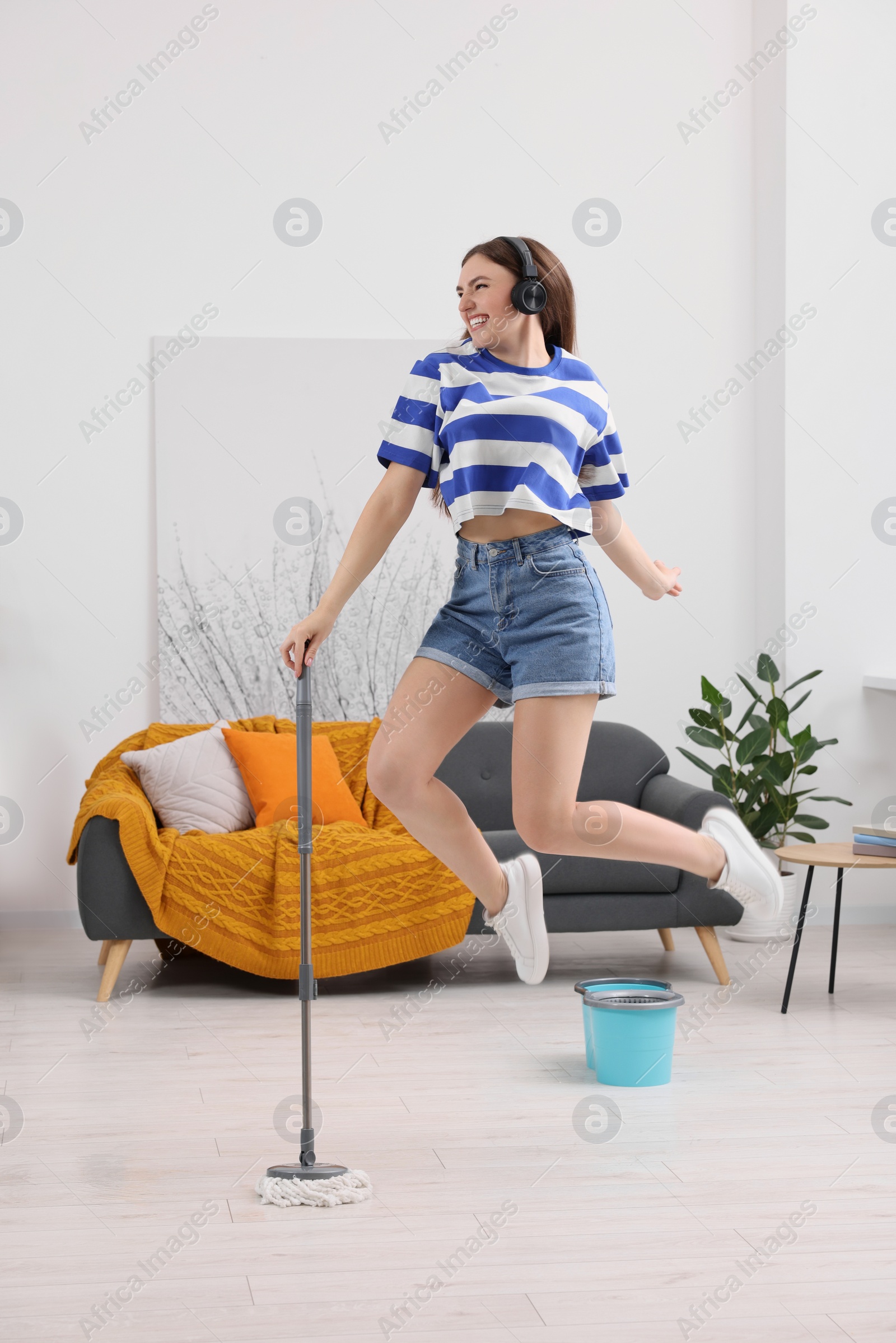 Photo of Enjoying cleaning. Happy woman in headphones jumping with mop at home