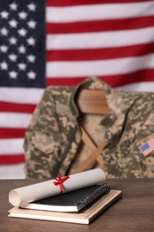 Photo of Notebooks and diploma on wooden table, chair with soldier uniform against flag of United States indoors. Military education