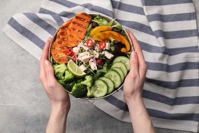 Photo of Woman holding bowl with many different vegetables and tofu at grey table, top view. Vegan diet