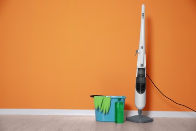 Modern steam mop, bucket with gloves and bottle of cleaning product on floor near orange wall, space for text