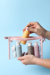 Cosmetic travel kit. Woman putting small bottle with personal care product into plastic bag against light blue background, closeup