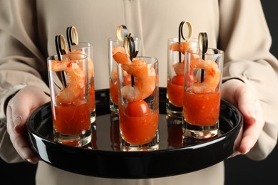 Photo of Woman holding tasty canapes with shrimps, tomatoes and sauce in shot glasses on black background, closeup