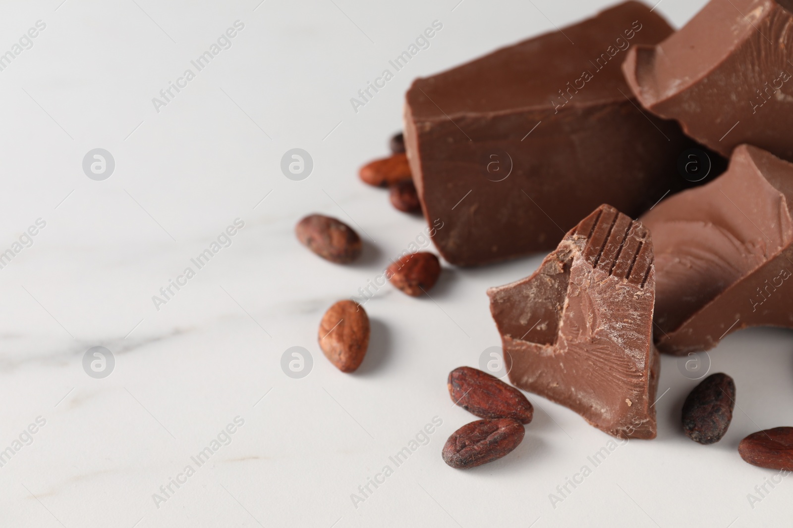 Photo of Pieces of tasty milk chocolate and cocoa beans on white marble table, closeup. Space for text