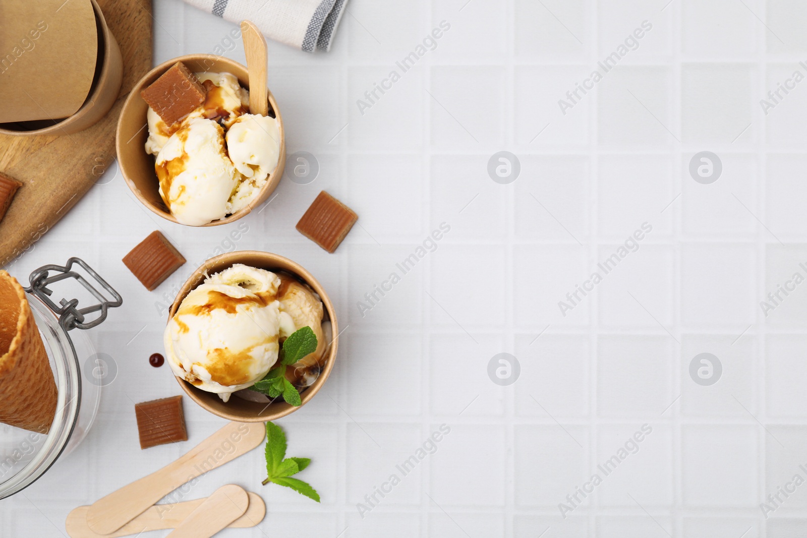 Photo of Scoops of ice cream with caramel sauce, mint leaves and candies on white tiled table, flat lay. Space for text