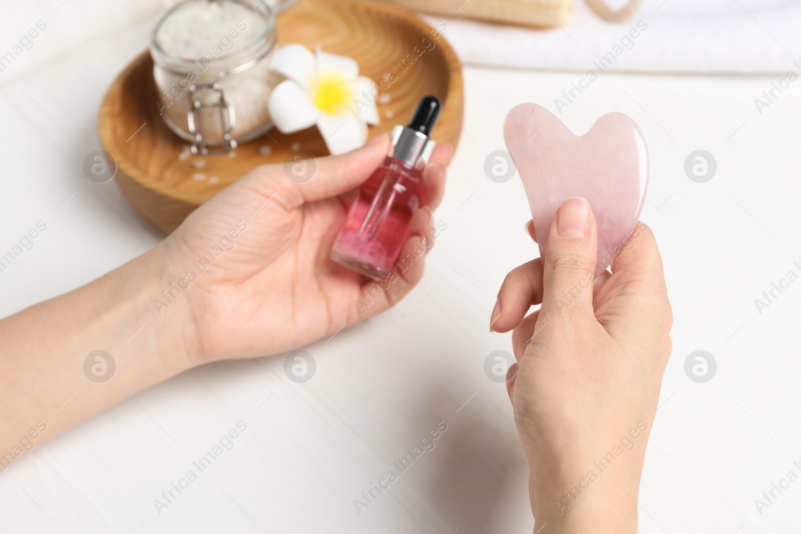 Photo of Woman holding gua sha tool and cosmetic at white wooden table, closeup