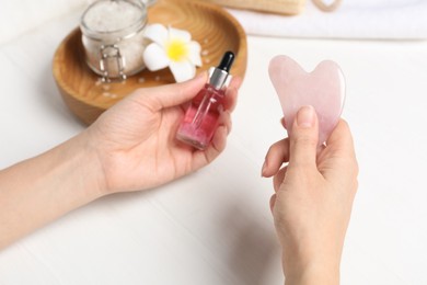 Photo of Woman holding gua sha tool and cosmetic at white wooden table, closeup