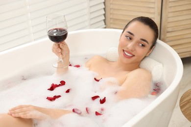 Photo of Happy woman with glass of wine taking bath in tub with foam and rose petals indoors