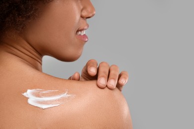 Young woman applying body cream onto back on grey background, closeup. Space for text