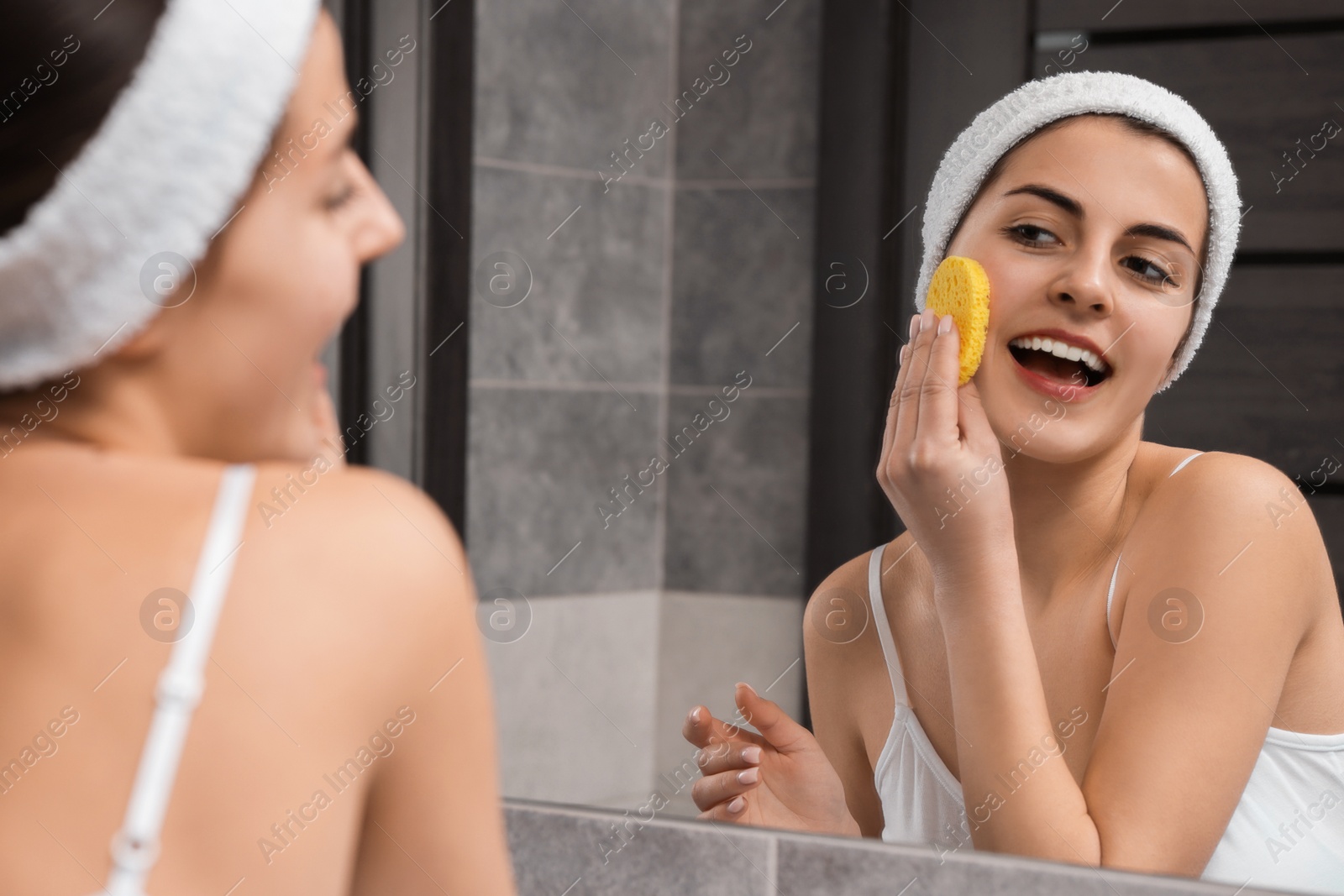 Photo of Young woman with headband washing her face using sponge near mirror in bathroom