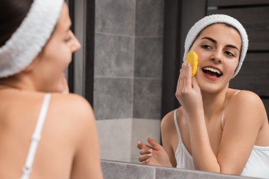 Young woman with headband washing her face using sponge near mirror in bathroom