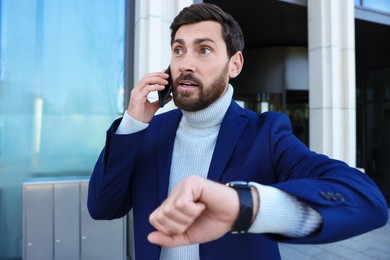 Photo of Emotional businessman talking on smartphone near building. Being late concept